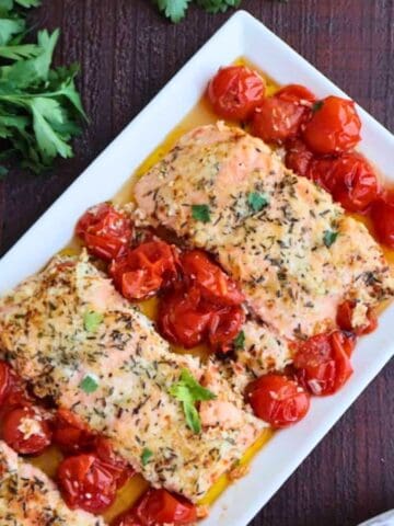 A white plate of 3 salmon filets surrounded by tomatoes.