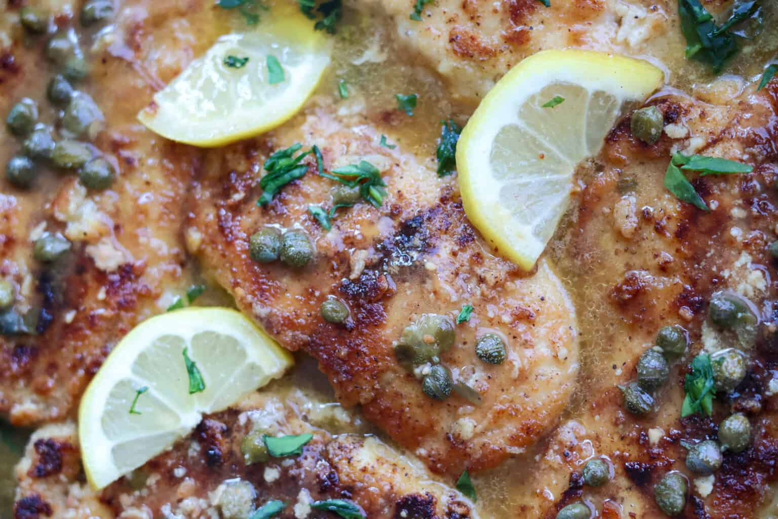 A close-up shot of chicken piccata. The dish features golden-brown, pan-fried chicken breasts garnished with lemon slices, capers, and chopped parsley, all topped with a light, buttery sauce. The chicken looks tender and well-seasoned.