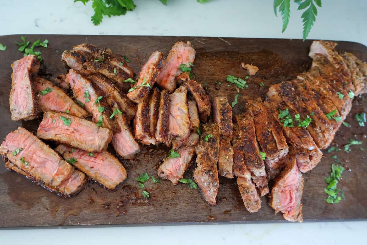 New York Strip steak slices on a brown cutting board with green garnish.