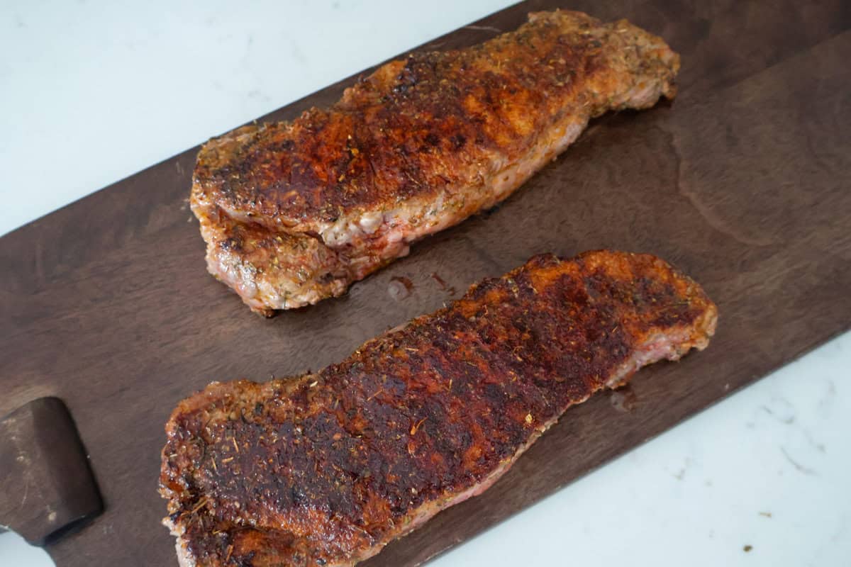 Two New York Strip Steaks finished cooking on a brown cutting board.