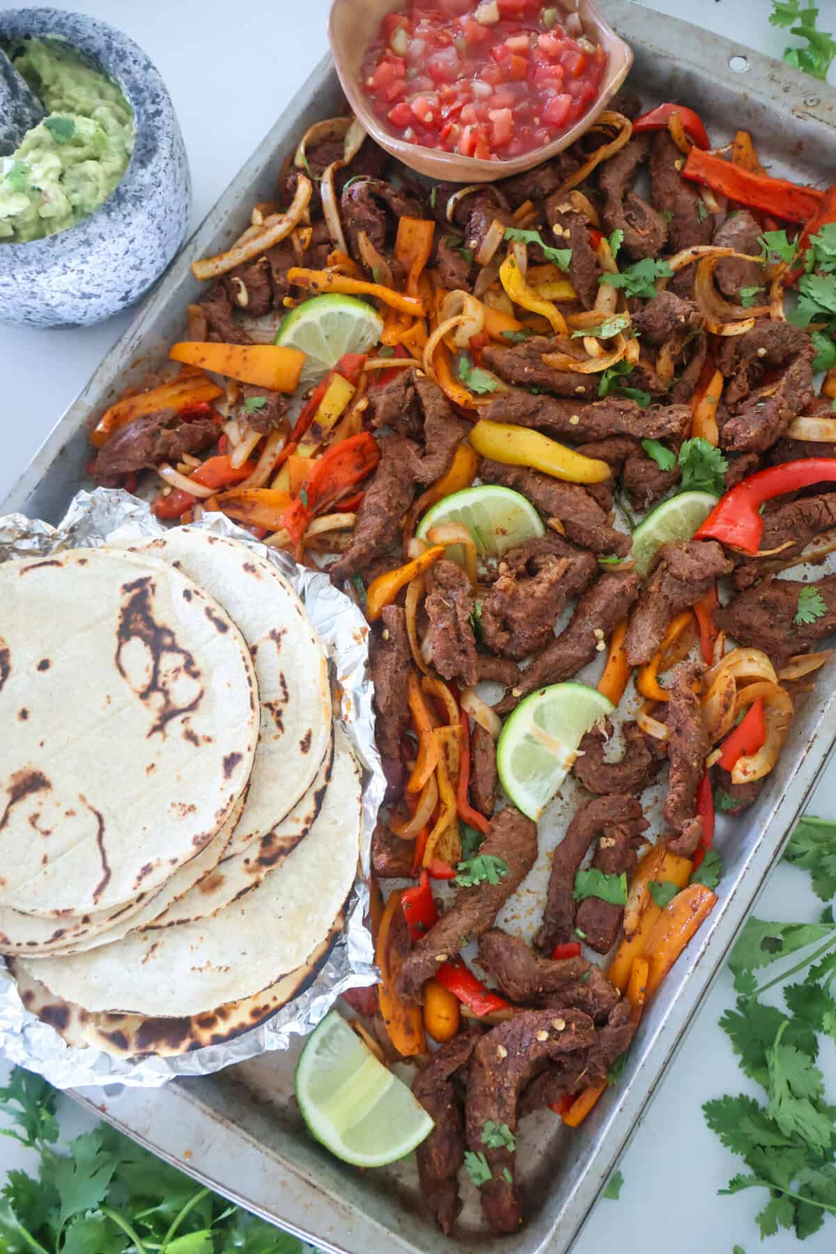 A sheet pan of Steak Fajitas with tortillas and salsa.
