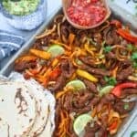 A sheet pan of Steak Fajitas with tortillas and salsa.