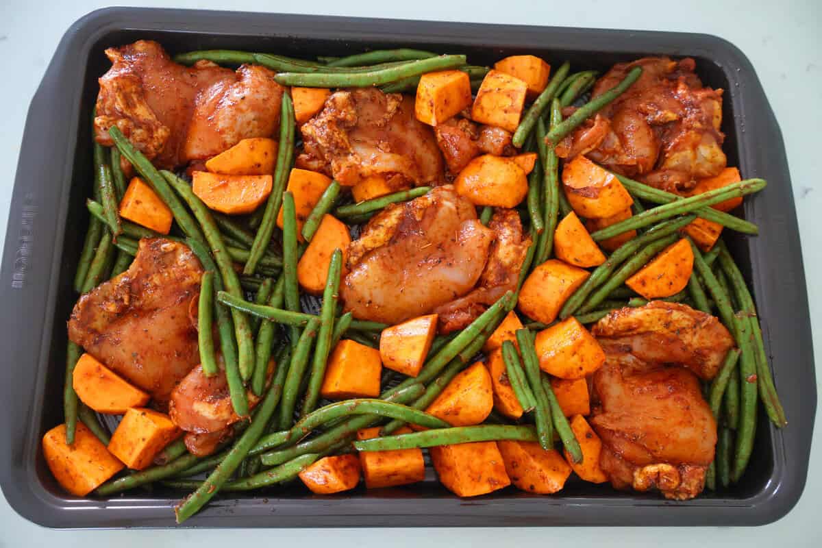 A sheet pan of raw chicken thighs, green beans and sweet potatoes.