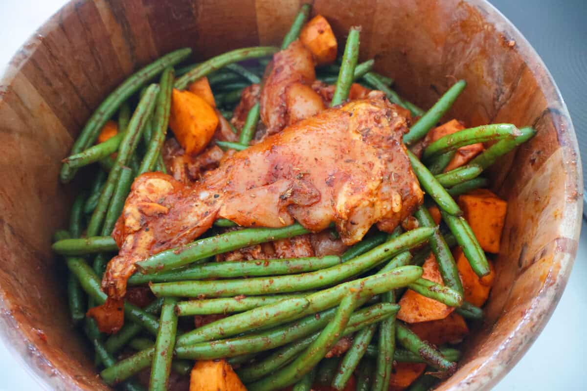 A wood bowl with raw chicken thighs, green beans and sweet potatoes.