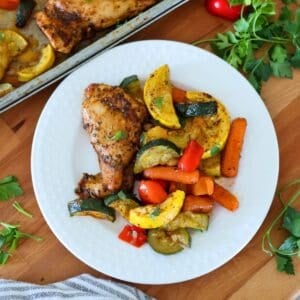 A white plate of sheet pan chicken and vegetables on a cutting board.