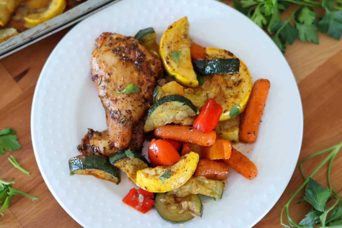 A white plate of sheet pan chicken and vegetables on a cutting board.