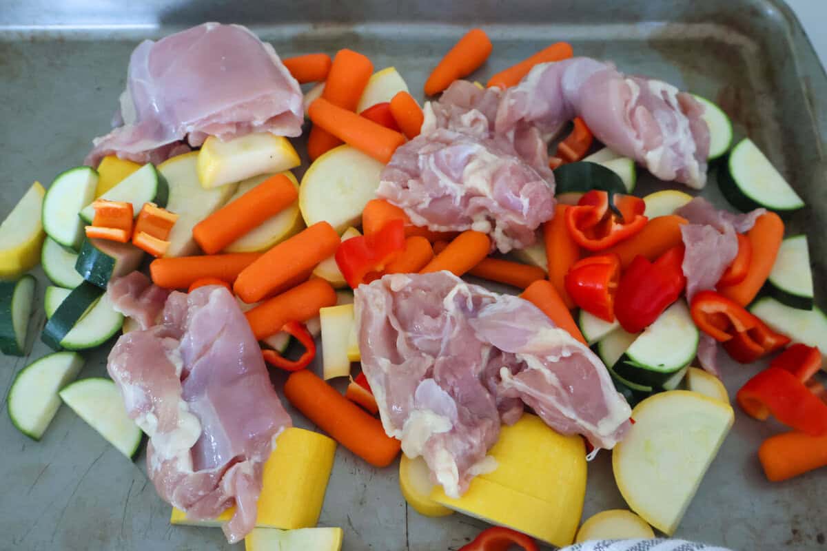 Raw chicken and zucchini and carrots on a baking sheet.
