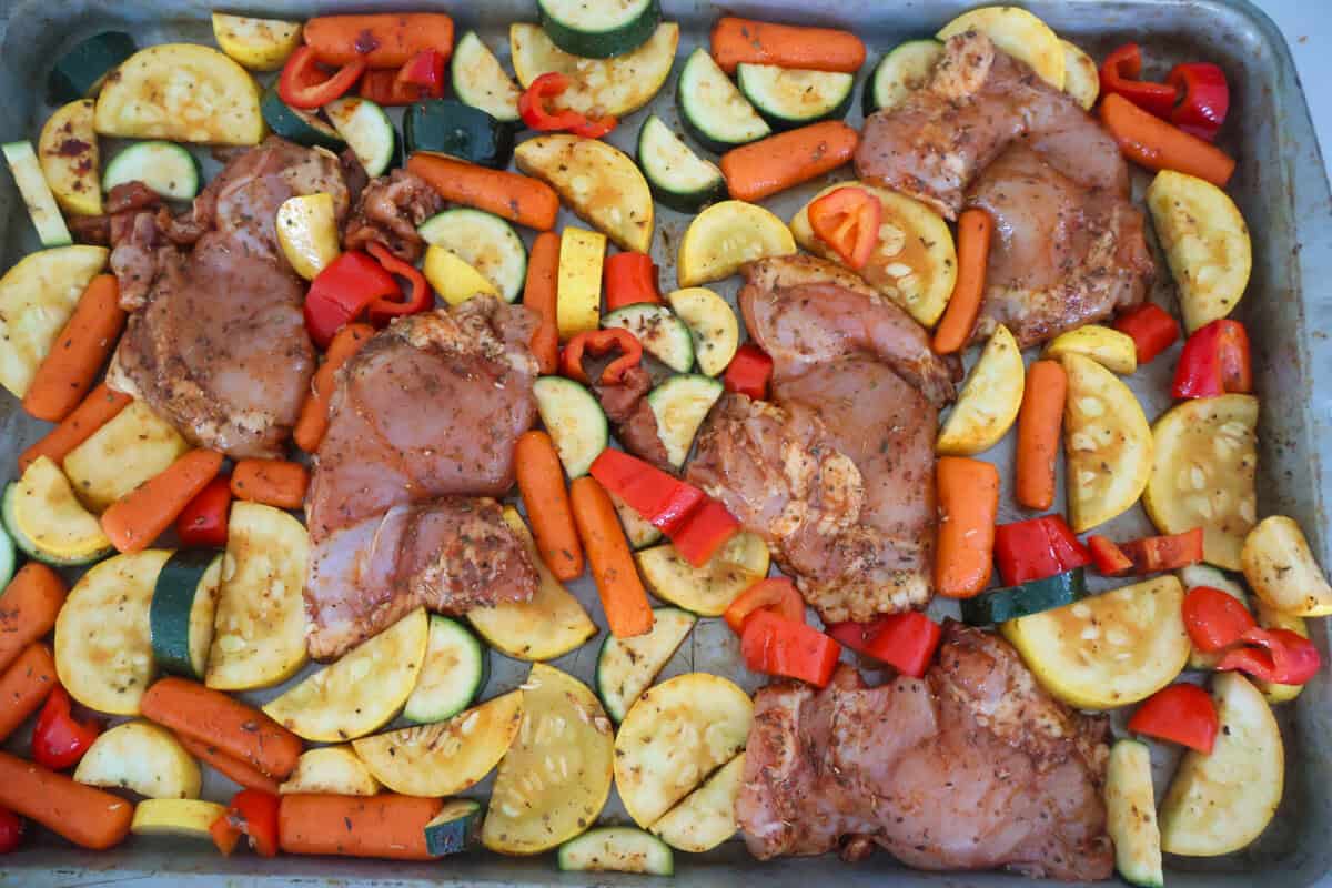 Raw chicken and zucchini and carrots with seasonings on a baking sheet.