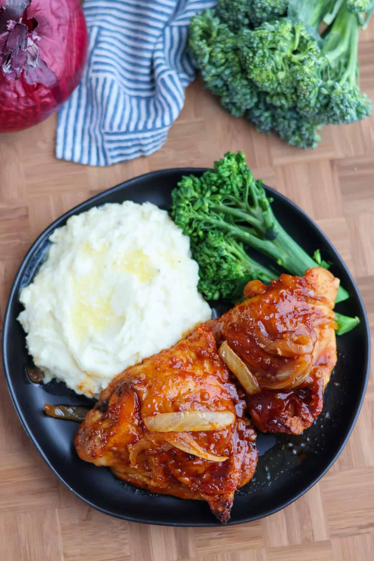 Baked BBQ chicken thighs plated with mashed potatoes and broccolini on a black plate