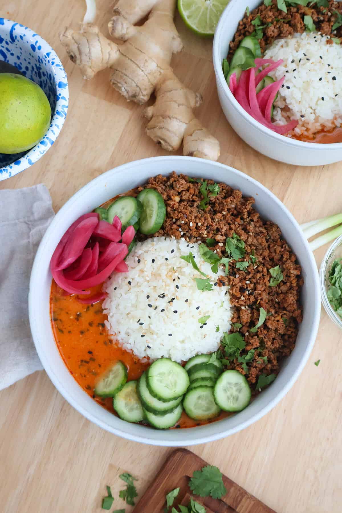 Crispy beef broth bowl topped with sesame seeds, sliced cucumbers, and pickled red onions next to fresh ginger and lime
