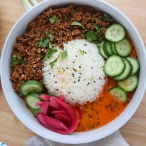 Overhead view of a crispy beef broth bowl with seasoned ground beef, white rice, red curry coconut broth, cucumbers, and pickled red onions