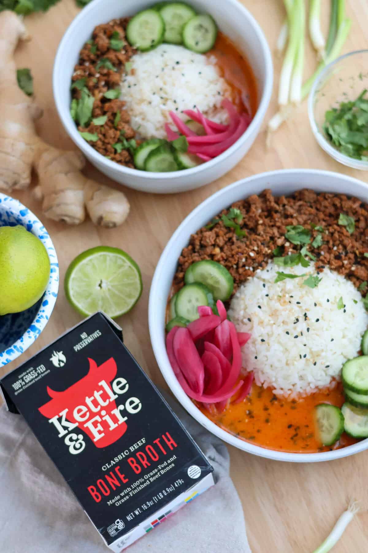 Two crispy beef broth bowls next to a carton of Kettle & Fire Classic Beef Bone Broth, fresh ginger, and limes